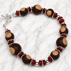 May include: A beaded bracelet featuring dark brown and tan beads, interspersed with red and silver beads. The bracelet has a toggle clasp. The beads are arranged in a repeating pattern, creating a unique and eye-catching accessory.