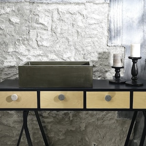 Pode incluir: Uma penteadeira preta com três gavetas e uma pia de concreto. A penteadeira está sobre pernas de metal preto e tem uma parede de pedra rústica ao fundo. Há dois castiçais pretos com velas brancas sobre a penteadeira.