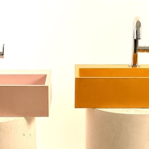 Può includere: Due lavandini rettangolari, uno rosa e uno giallo, ciascuno con un rubinetto cromato. I lavandini sono montati su piedistalli cilindrici bianchi su sfondo bianco. I lavandini hanno un design moderno e minimalista.