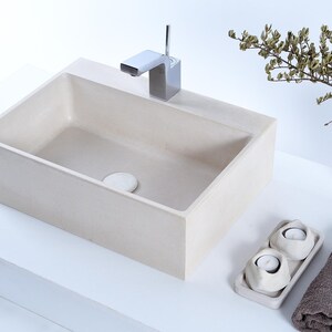 May include: A rectangular off-white concrete bathroom sink with a chrome faucet, a matching drain cover, a tall concrete vase with dried branches, a rectangular tray holding two white tea light candle holders, and a folded dark gray towel.