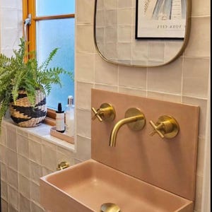 May include: A light pink bathroom sink with a gold faucet and drain. The sink is mounted on a wall with a light pink tile backsplash. There are two gold knobs on the wall above the faucet.