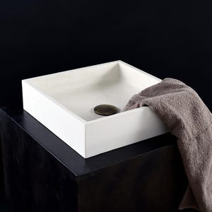 May include: A white square concrete vessel sink with a brass drain cover. The sink is sitting on a black pedestal and a brown towel is draped over the side.