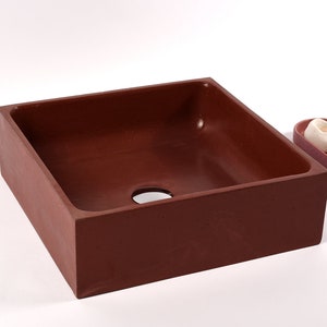 May include: A square, brown concrete vessel sink with a drain hole in the center. The sink is on a white surface.