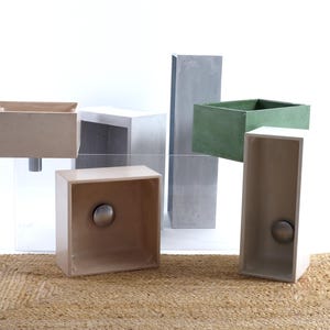 Op de afbeelding: Vijf betonnen badkamer wastafels in verschillende maten en vormen. De wastafels zijn gerangschikt op een lichtbruin kleed. De wastafels zijn allemaal lichtbeige van kleur, behalve één die lichtgroen is.