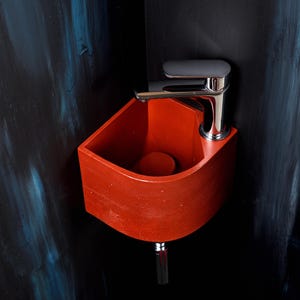 Può includere: Un moderno lavabo rosso a parete con un rubinetto cromato. Il lavabo ha un design curvo ed è posto su uno sfondo scuro con pennellate blu e nere. Il rubinetto è posizionato sopra il bacino del lavabo.