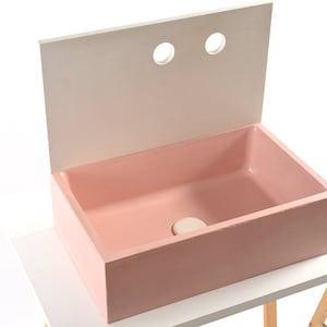 Puede incluir: Un lavabo de hormigón rosa con un salpicadero blanco y dos agujeros. El lavabo está sobre una mesa blanca con patas de madera.