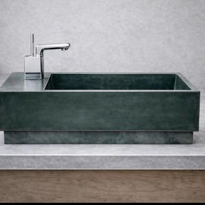 Op de afbeelding: Een rechthoekige, donkergroene betonnen wastafel met een chromen kraan. De wastafel staat op een grijs aanrechtblad met een houten onderstel. De kraan is modern en hoekig. De uitstraling is minimalistisch en modern.