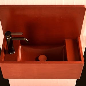 May include: A contemporary, terracotta-coloured concrete sink with an integrated backsplash and a rectangular basin. It has a black tap and a small, round drain cover. The sink is wall-mounted.