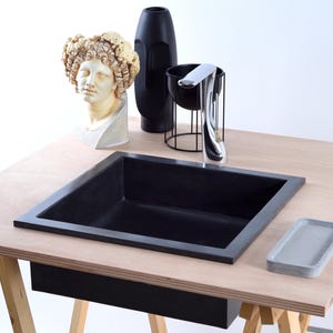 May include: A black concrete sink with a square shape sits on a wooden table with a light wood finish. A chrome faucet is mounted on the table behind the sink. A small gray rectangular soap dish is on the table to the right of the sink.