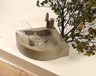May include: A gray concrete corner sink with a curved front edge and a brushed metal faucet. Water is splashing inside the basin, creating a dynamic movement. The sink is designed for corner installation and features a smooth, solid construction.