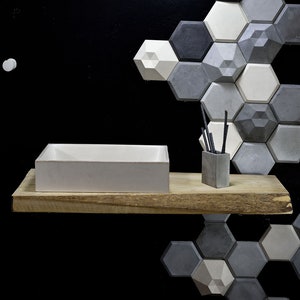 May include: A white rectangular concrete sink sits on a wooden shelf with a small concrete container holding black sticks. A wall behind the shelf is covered with a geometric pattern of gray and white concrete hexagonal tiles.