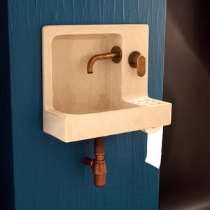 May include: A wall-mounted beige concrete sink featuring a curved copper faucet and a matching copper handle. The basin includes an integrated soap dish with drainage holes and a built-in toilet paper holder underneath, with a copper drain pipe below.
