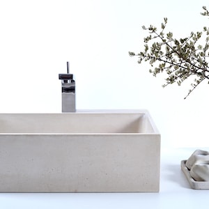 Könnte beinhalten: Ein rechteckiges Waschbecken aus Beton mit einem verchromten Wasserhahn. Das Waschbecken steht auf einer weißen Oberfläche mit einem braunen Handtuch und einem weißen Dekorationsgegenstand.