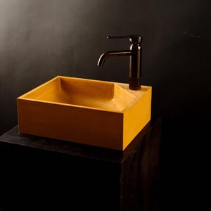 Könnte beinhalten: Ein rechteckiges, senfgelbes Waschbecken mit glatter, matter Oberfläche und einer einzelnen schwarzen Metallarmatur auf der rechten Seite. Das Waschbecken zeichnet sich durch klare, scharfe Kanten und ein tiefes Becken aus, das einen Kontrast bildet.