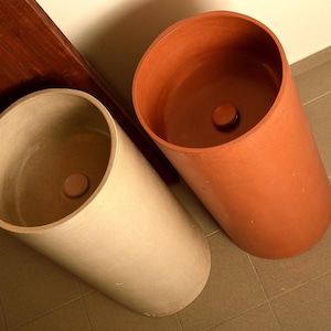 Könnte beinhalten: Zwei zylindrische Standwaschbecken, eines in hellbeiger Betonoptik und das andere in terrakotta-orangem Farbton. Beide Becken verfügen über ein glattes, tiefes Inneres mit einer passenden runden Abflussabdeckung in der Mitte des Bodens.