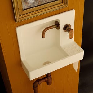 May include: A small, white, wall-mounted sink with a copper faucet and fixtures. The sink has a rectangular basin and a small soap dish. The plumbing is copper. The sink is mounted on a textured, yellow-orange wall.