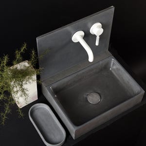 May include: A modern, dark gray concrete bathroom sink with a rectangular basin and a matching backsplash. White faucet and handle fixtures are mounted on the backsplash. An oval soap dish and a potted plant are next to the sink.