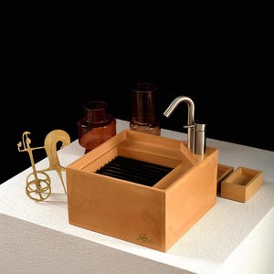 Könnte beinhalten: Ein quadratisches, hellorangefarbenes Waschbecken mit einem Armatur aus gebürstetem Metall, einem schwarzen Lamelleneinsatz und einer Signatur auf der Vorderseite. Daneben stehen zwei bernsteinfarbene Glaszylinder, eine goldene Streitwagenskulptur und zwei kleine rechteckige Schalen.
