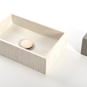 Könnte beinhalten: Ein weißes rechteckiges Betonwaschbecken mit einer strukturierten Oberfläche und einem weißen Abflussstopfen. Das Waschbecken steht auf einer weißen Oberfläche. Zwei graue Betonblöcke mit Löchern oben befinden sich im Hintergrund.