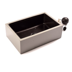 May include: A rectangular basin with a smooth light gray exterior and a deep black interior. The basin features a central drain hole at the bottom. Positioned near the top right corner is a small, round black perfume atomizer with a silver nozzle.
