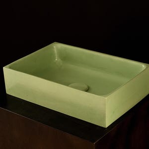 Puede incluir: Un lavabo rectangular de cerámica verde claro con un agujero de desagüe en el centro. El lavabo está sobre una superficie marrón oscuro.