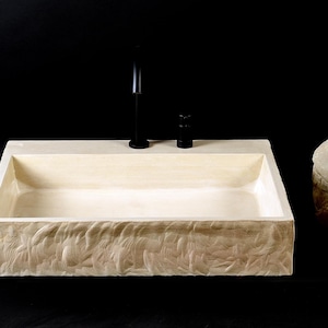 May include: A rectangular stone sink with a smooth interior basin and a textured, chiseled exterior surface. The sink features a flat rear ledge with a tall, slender black faucet and a matching black control knob, accompanied by two natural stones.