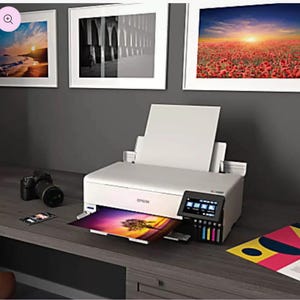 May include: A white Epson printer with a color display and ink cartridges. A printed photo is emerging from the printer. A digital camera and a smartphone are on the desk. Framed art pieces hang on the wall.