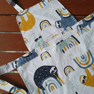 May include: A white apron featuring a colourful rainbow and sloth design. The apron has adjustable straps and a large pocket on the front.