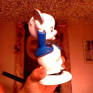 May include: A white ceramic figurine of a cartoon character with a blue and white outfit. The character is holding a round object in its right hand and has a black bow tie.