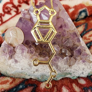 以下が含まれることがあります： 分子構造のデザインが施された、ゴールドカラーのペンダントネックレス。ペンダントは紫のアメジストクリスタル上に展示されており、1976年のペニーがスケールとして表示されています。ネックレスチェーンもゴールドです。