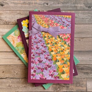 May include: Three notebooks with different floral patterns and a purple ribbon tied around the top notebook. The top notebook has a floral pattern of purple, pink, orange, and yellow tulips.