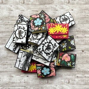 May include: A collection of small, decorative boxes with various floral and geometric patterns. The boxes are made of black and white paper with accents of red, yellow, and blue. The boxes are arranged in a pile on a white wooden surface.