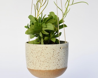 Puede incluir: Una maceta colgante con una maceta de cerámica blanca moteada y una base marrón, suspendida por cuerdas naturales. La maceta contiene una planta suculenta verde con múltiples tallos y hojas, sobre un fondo blanco.