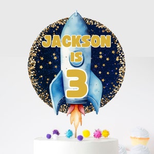 Op de afbeelding: Een blauwe en gouden rakettaarttopper met de tekst "JACKSON IS 3" op een blauwe en gouden glitterachtergrond.