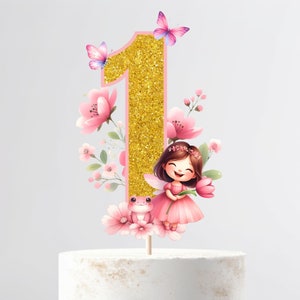 May include: A gold glitter number one cake topper with pink details, butterflies, and flowers. A smiling fairy in a pink dress and a small pink frog are also featured. This decorative topper is perfect for a first birthday celebration.