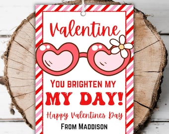 May include: A Valentine's Day gift tag with red and pink stripes. It features a cartoon image of heart-shaped sunglasses with a flower. The text reads "Valentine You Brighten My Day! Happy Valentines Day From Maddison".
