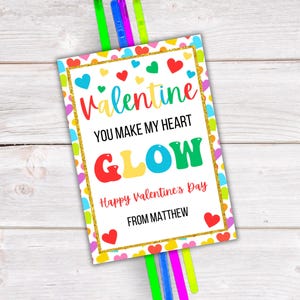 Op de afbeelding: Een Valentijnskaart met een witte achtergrond en een gouden glitterrand. De kaart heeft een kleurrijk hartontwerp en de tekst "Valentine You Make My Heart Glow Happy Valentine's Day From Matthew".