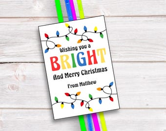 May include: A white Christmas card with the message "Wishing you a BRIGHT and Merry Christmas From Matthew" is decorated with colorful string lights. The card is paired with green, blue, pink, and yellow glow sticks, all set against a light wood background.