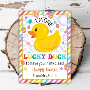 May include: A rectangular tag with a yellow rubber duck illustration, bubbles, and Easter eggs. Text includes "I'M ONE", "LUCKY DUCK", "Happy Easter", and "From Mrs. Smith". The tag has a rainbow striped border.
