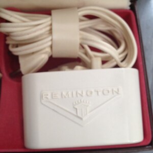 May include: White electric shaver charging base with a white cord wrapped around it. The base has the word "REMINGTON" printed on it in black letters.