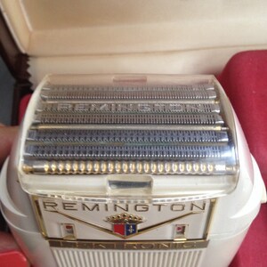 May include: White electric razor with a clear plastic cover over the blades. The razor has the word "REMINGTON" printed on the front in gold letters. The razor also has a gold crest with a fleur-de-lis.