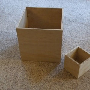 May include: Two wooden boxes, one larger than the other, are shown on a light brown carpet. Both boxes are open and empty.