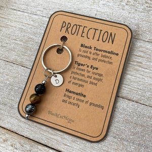 May include: A brown card with the word "PROTECTION" printed in black. The card lists the properties of Black Tourmaline, Tiger's Eye, and Hematite. A silver keychain with a letter "D" charm and three gemstone beads: black, brown, and grey.
