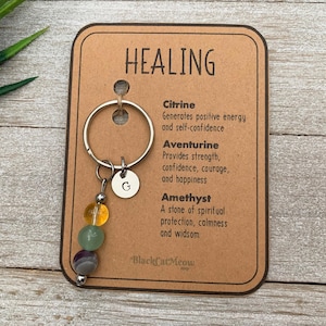 May include: A silver keychain with a charm featuring three gemstone beads: citrine, aventurine, and amethyst. The charm is attached to a silver key ring with a letter 'G' charm. A card with the text "HEALING" and descriptions of the healing properties of each gemstone is included.
