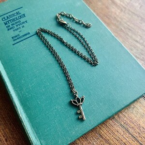 Puede incluir: Un collar de metal en tono bronce con un pequeño colgante en forma de llave con una parte superior similar a una corona. La cadena es delicada e incluye una sección de eslabones ajustable. El colgante tiene un acabado oscuro y antiguo.