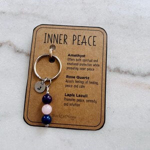 Puede incluir: Un llavero sobre una tarjeta rectangular con la etiqueta INNER PEACE, que incluye una anilla de metal plateado, un peque&ntilde;o dije circular grabado con la letra J y tres cuentas en azul oscuro, rosa claro y azul oscuro, con texto sobre amatista, cuarzo rosa y lapisl&aacute;zuli.