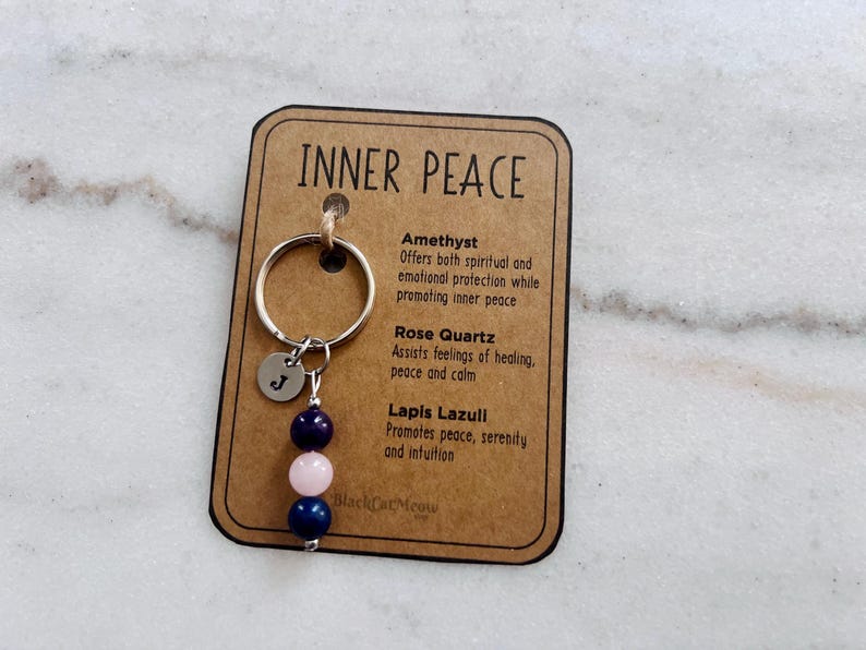 Puede incluir: Un llavero sobre una tarjeta rectangular con la etiqueta INNER PEACE, que incluye una anilla de metal plateado, un peque&ntilde;o dije circular grabado con la letra J y tres cuentas en azul oscuro, rosa claro y azul oscuro, con texto sobre amatista, cuarzo rosa y lapisl&aacute;zuli.