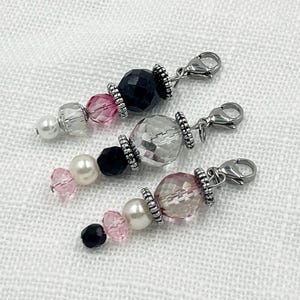 May include: Three beaded charms with lobster claw clasps. Each charm features a combination of black, pink, clear, and pearl-colored beads. The beads are separated by silver-colored metal accents. The charms are displayed on a white, textured surface.