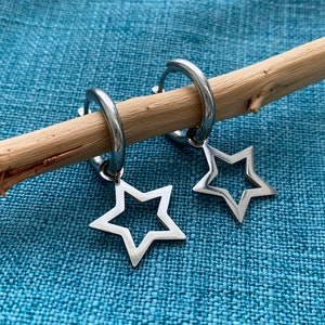 Può includere: Un paio di orecchini a cerchio argentati con ciondoli a forma di stella. Gli orecchini sono esposti su un bastoncino di legno, su uno sfondo blu strutturato. I ciondoli a stella hanno un design traforato.