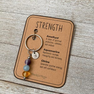 May include: A keychain with three gemstone beads: amethyst, aquamarine, and citrine. The keychain has a silver charm with the letter "N" and a silver key ring. The card attached to the keychain reads "STRENGTH" and lists the properties of each gemstone.